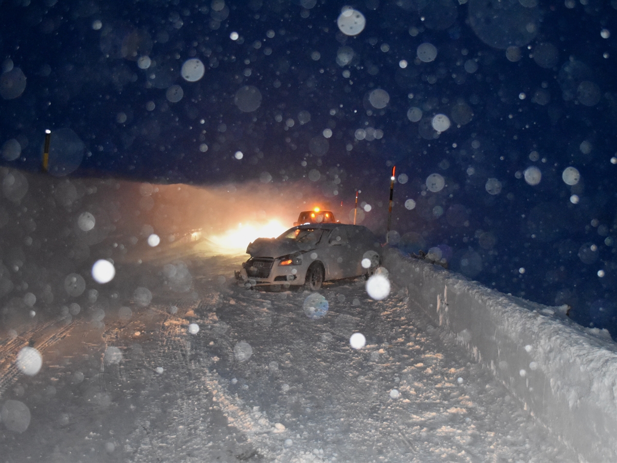 Bild in fortgeschrittener Abenddämmerung. Inmitten der beidseitig mit Schneewänden begrenzten Fahrbahn ein frontal stark beschädigtes Auto von schräg vorne gesehen.
