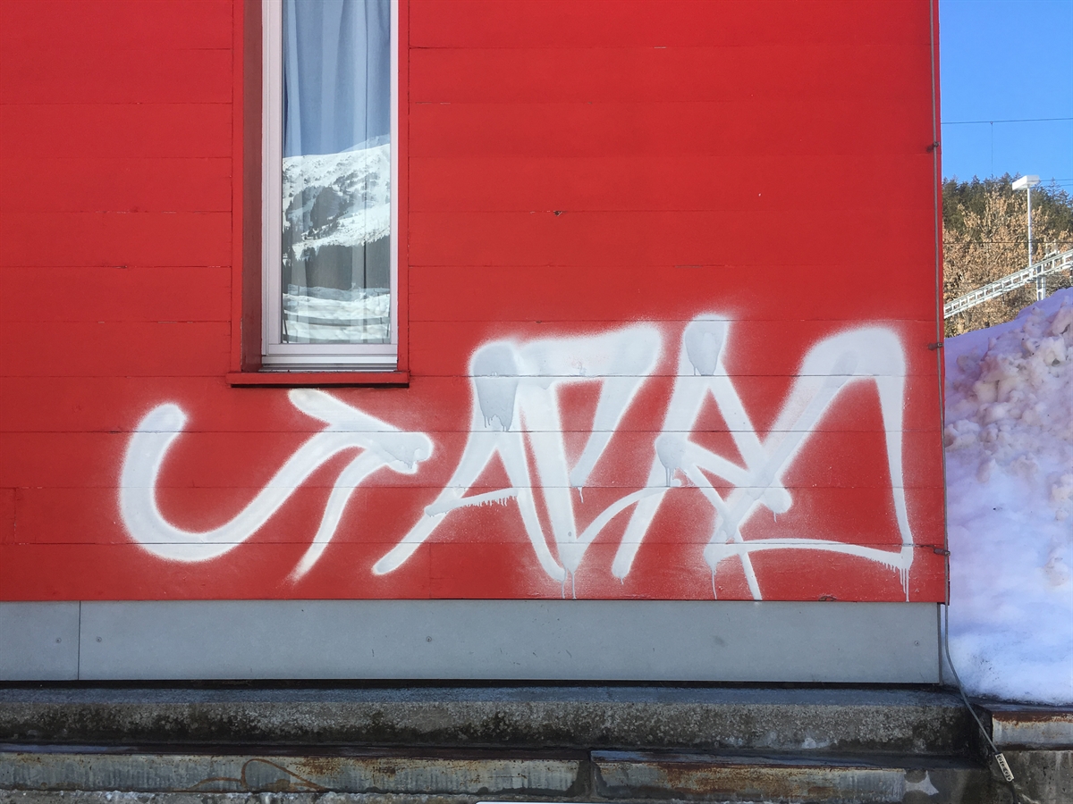 Ein mit weisser Farbe bespraytes rotes Gebäude. Die gesprayten Buchstaben sind als Wort erkennbar, welches Wort ist nicht ganz klar.