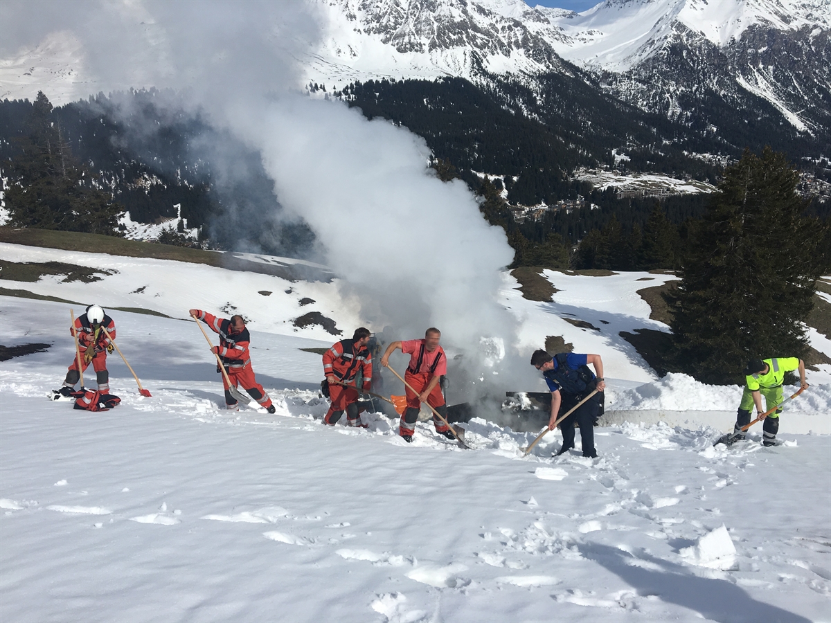 Feuerwehrleute, ein Polizist und der Fahrer schaufeln Schnee auf die in Brand geratene Schneefräste.