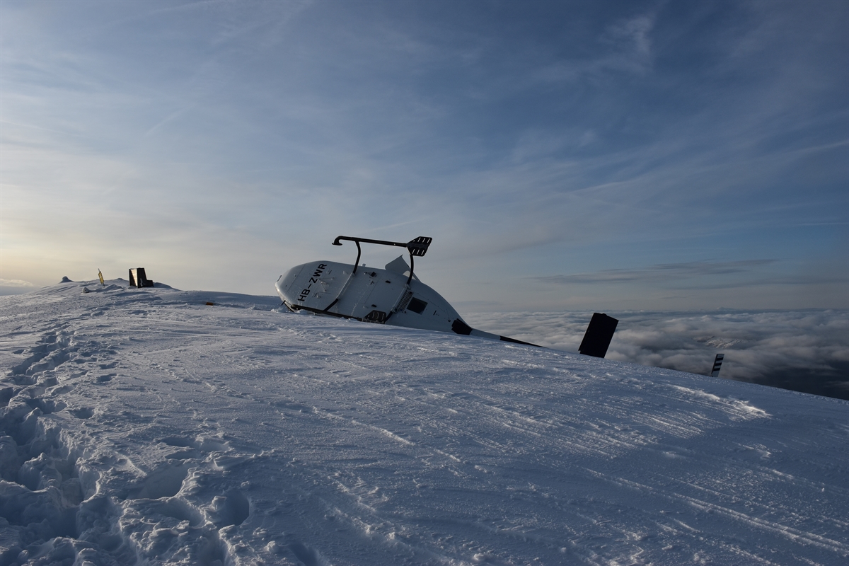 Der Helikopter liegt im Schnee auf einem Bergrücken auf der rechten Seite. Die Aufnahme zeigt die Unterseite des Helikopters. Im Hintergrund das Bergpanorama.