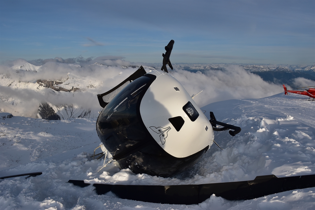 Der auf der rechten Seite im Schnee liegende Helikopter, rechts dahinter das Heck eines weiteren Helikopters sowie im Hintergrund das Bergpanorama. 