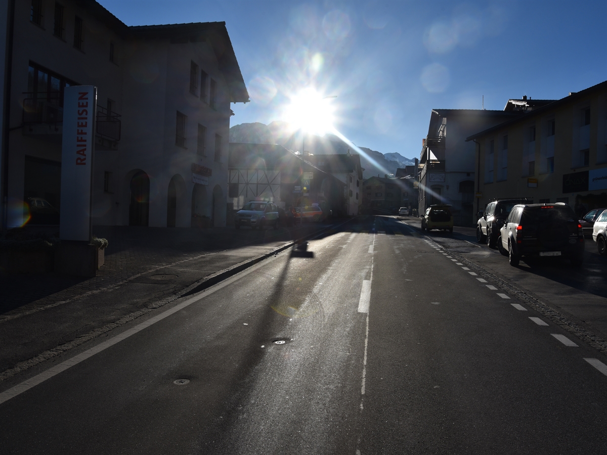 Die Italienische Strasse im Bereich der Post / Raiffeisenbank in Cazis. Die Sonne scheint auf die Fahrbahn.