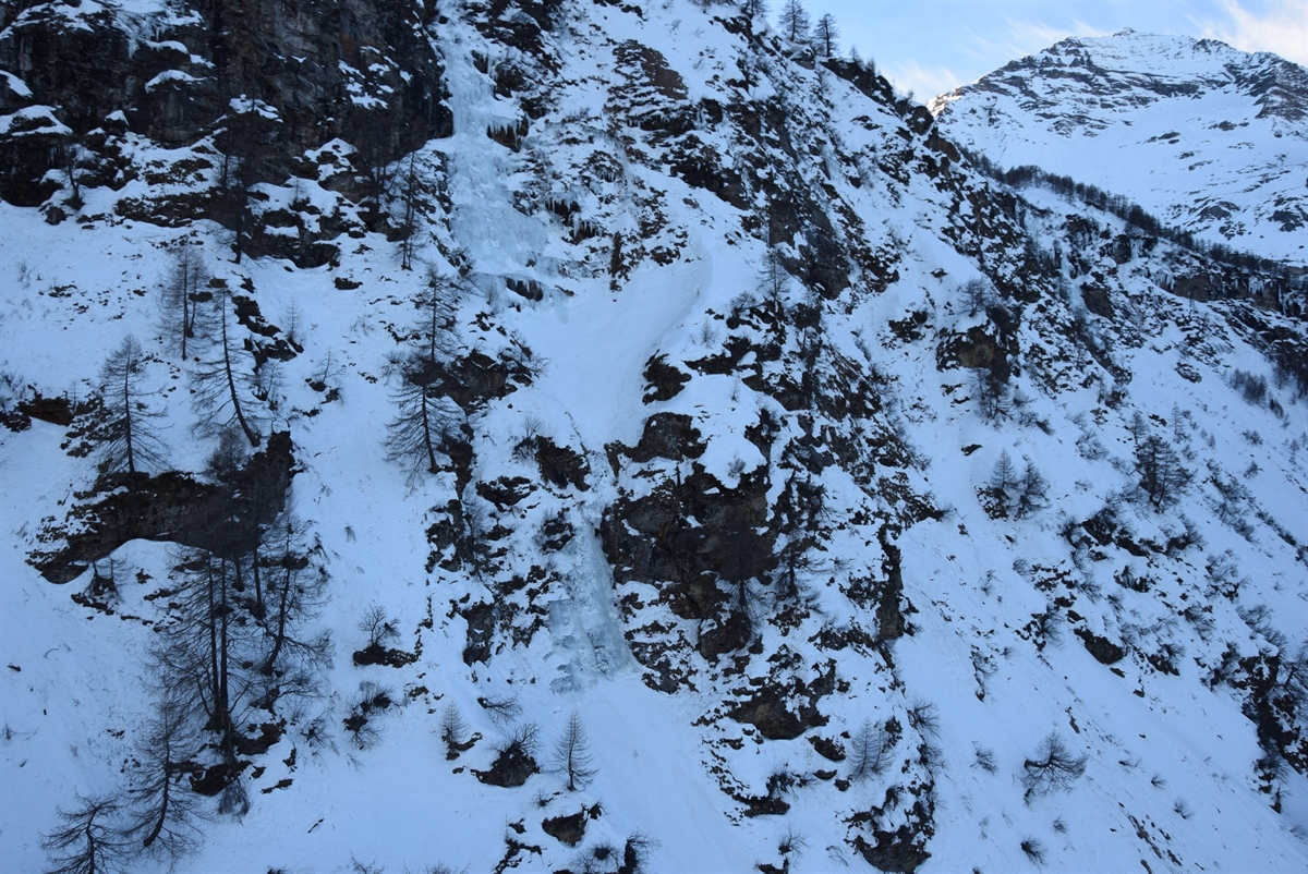 Stark abfallendes, verschneites und felsdurchsetztes Gelände. Inmitten davon eine beinahe durchgehende Eisfläche eines Wasserfalls. 