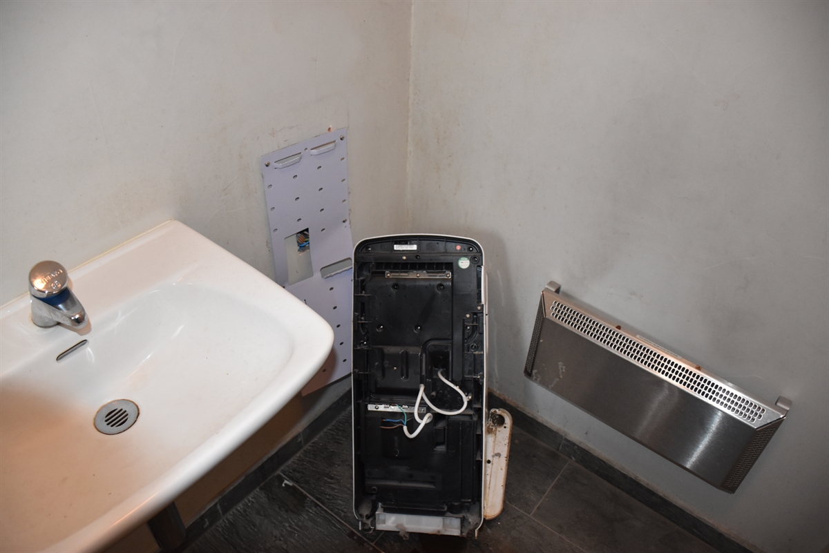 Ecke des Toilettenraums, links im Bild das Lavabo, rechts daneben am Boden der von der Wand gerissene Haartrockner.