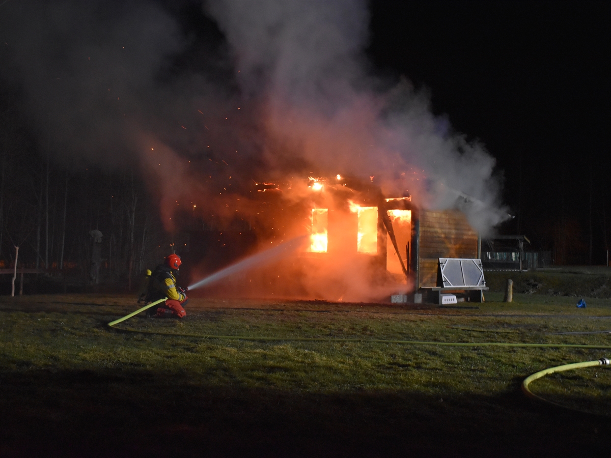 Ein Feuerwehrmann kniet vor dem Holzschuppen und spritzt Wasser in dieses. Flammen lodern aus den Gebäudeöffnungen hervor.