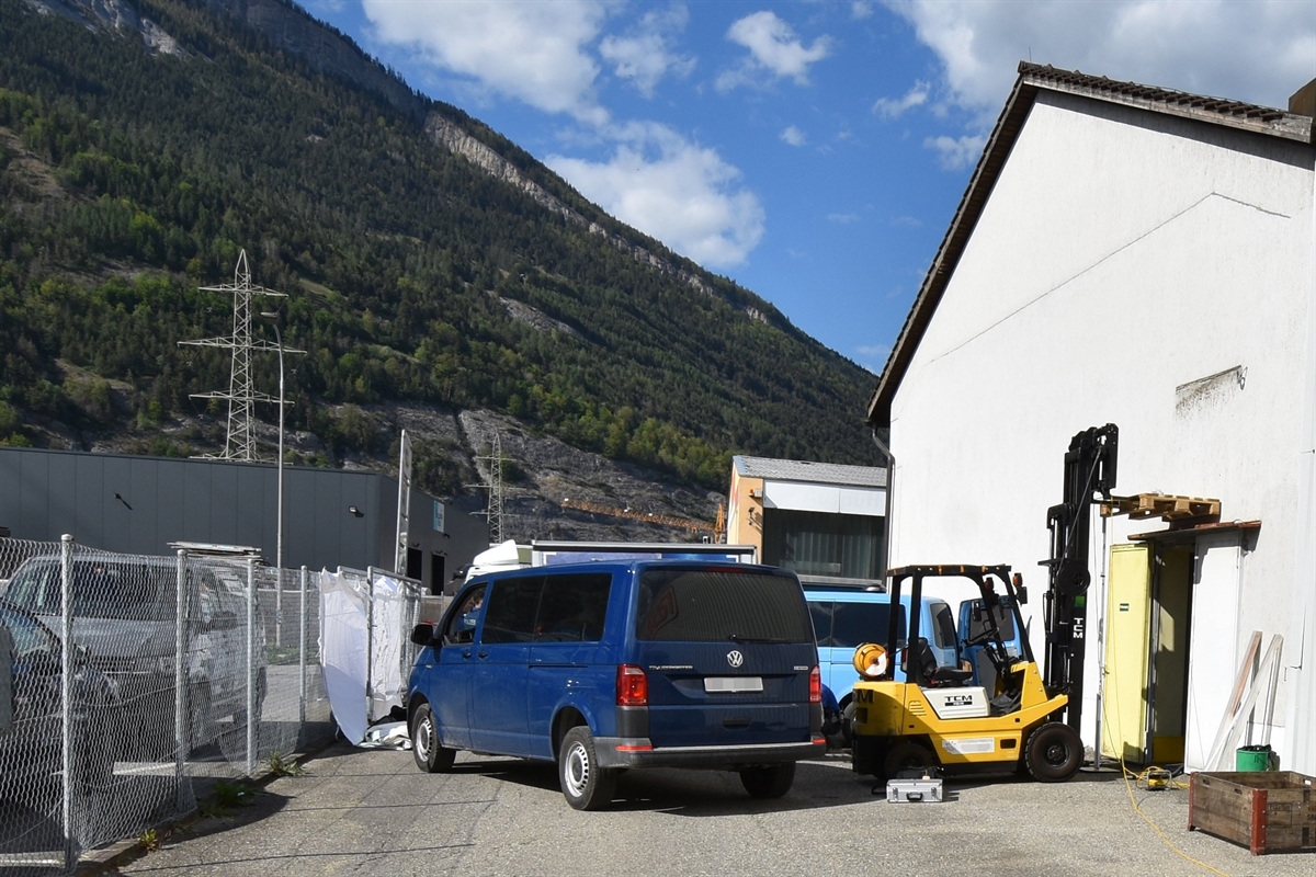 Ein Firmengelände mit Fahrzeugen, im Hintergrund der Calanda. Rechts ein Gebäude, an dessen linker Fassade ein Hubstabler mit angehobener Holzpalette. 