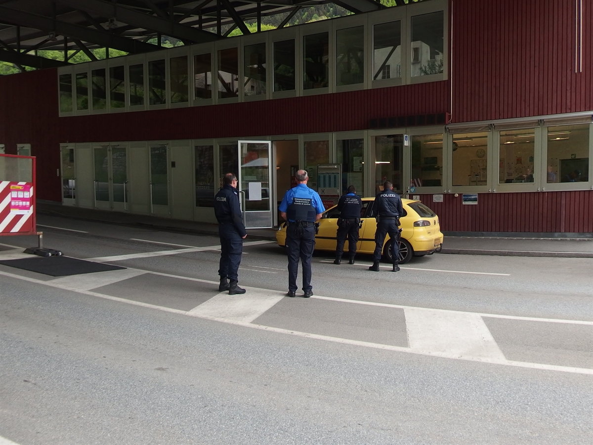 Von hinten total vier Mitarbeitende der Polizei und der EZV. Zwei kontrollieren ein gelbes Auto. Im Hintergrund das Gebäude der Grenzstation.