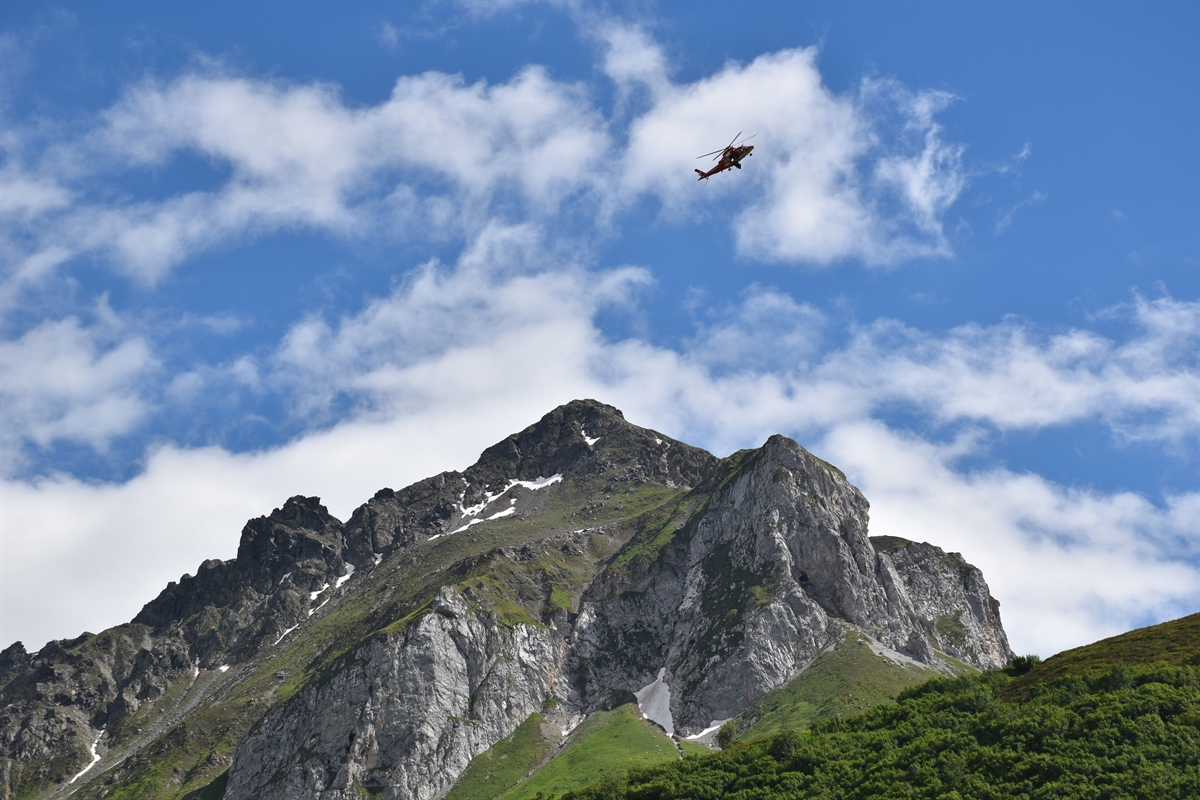 Oben mittig im Bild der fliegende Rega-Helikopter, blauer Himmel und einige Wolken im Hintergrund. In der unteren Bildhälfte ein Berg sowie ein unten links nach oben rechts zulaufender Bergrücken.
