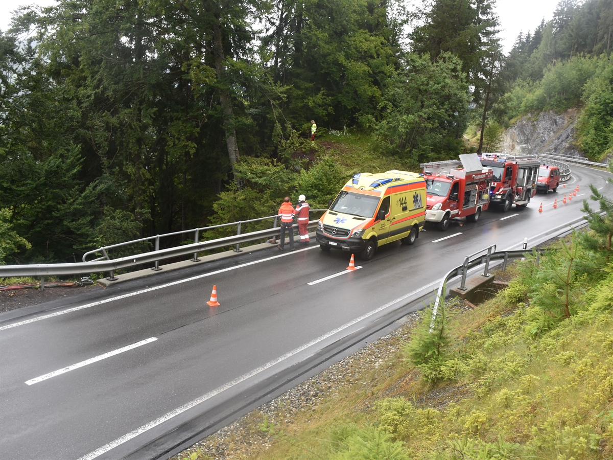 Die Strasse mit einem Ambulanz- und drei Feuerwehrfahrzeugen sowie Einsatzkräften, welche die Arbeiten im Tobel überwachen. 