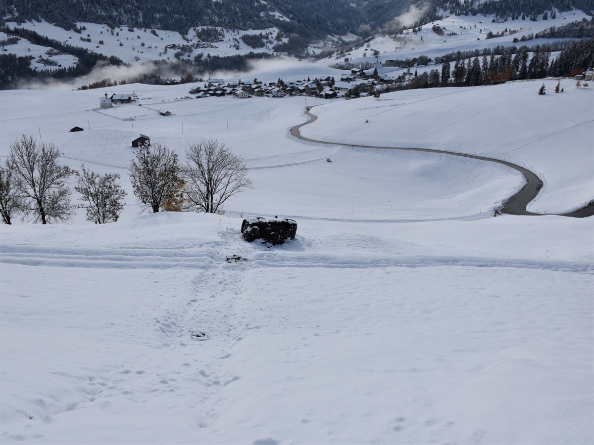 Aufnahme in das abfallende verschneite Wiesland. Inmitten diesem liegt auf der Fahrerseite das Auto. Im Hintergrund das Dorf Degen.