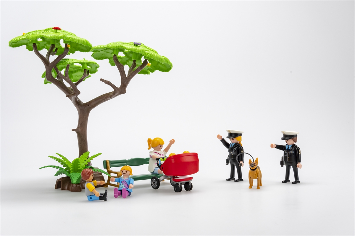 Eine Szenerie mit Playmobil-Figuren. Rechts eine Polizistin und ein Polizist mit Polizeihund, welche nach links zu einer auf einer Bank sitzenden Frau mit Kinderwagen blicken. Sie winken einander zu. Links vor der Bank zwei am Boden spielende Kinder.