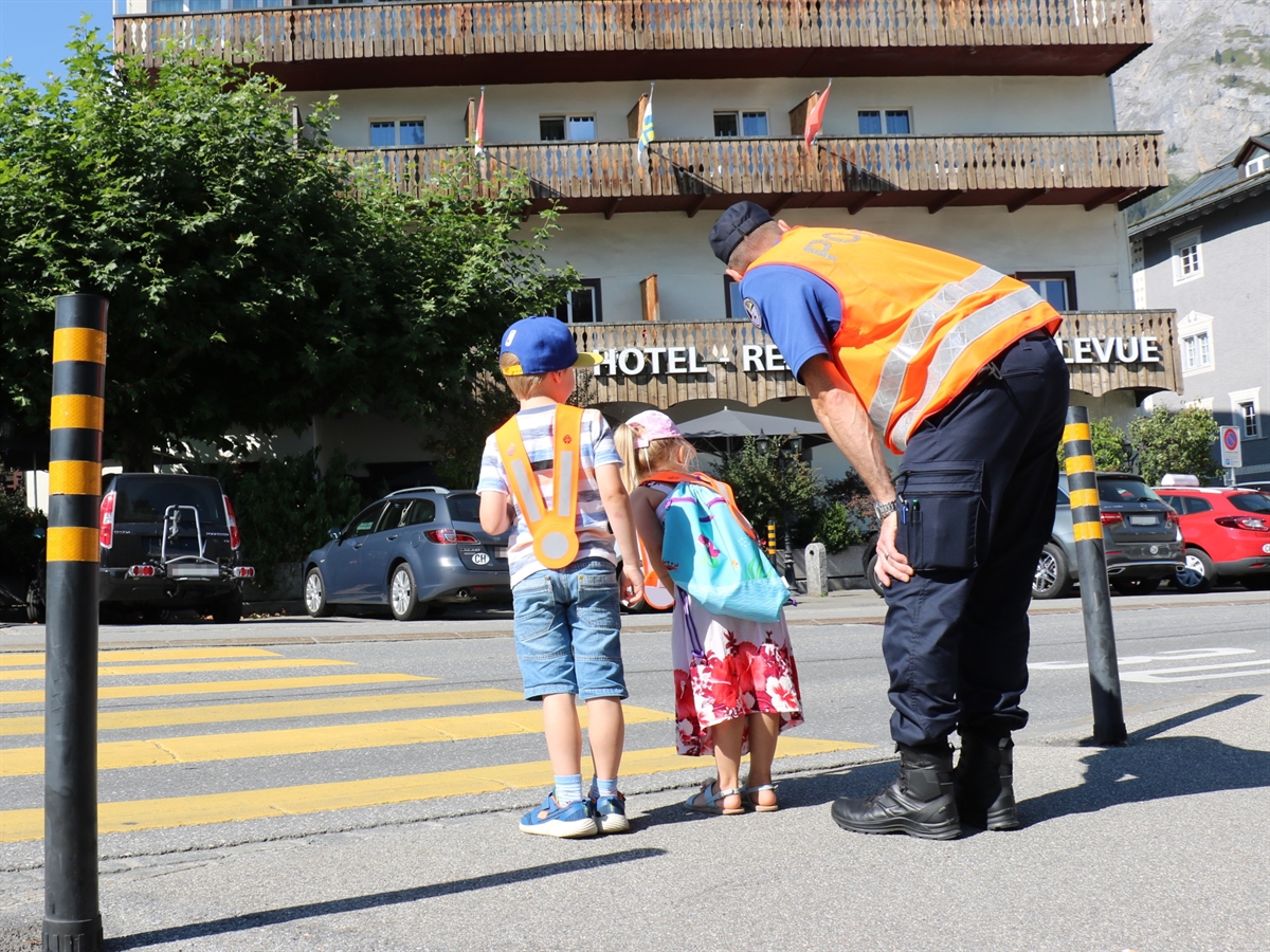 Zwei Kinder an einem Fussgängerstreifen und nach rechts auf die Strasse schauen. Rechts von ihnen ein Polizist, welcher sich zu ihnen hinunterbückt.