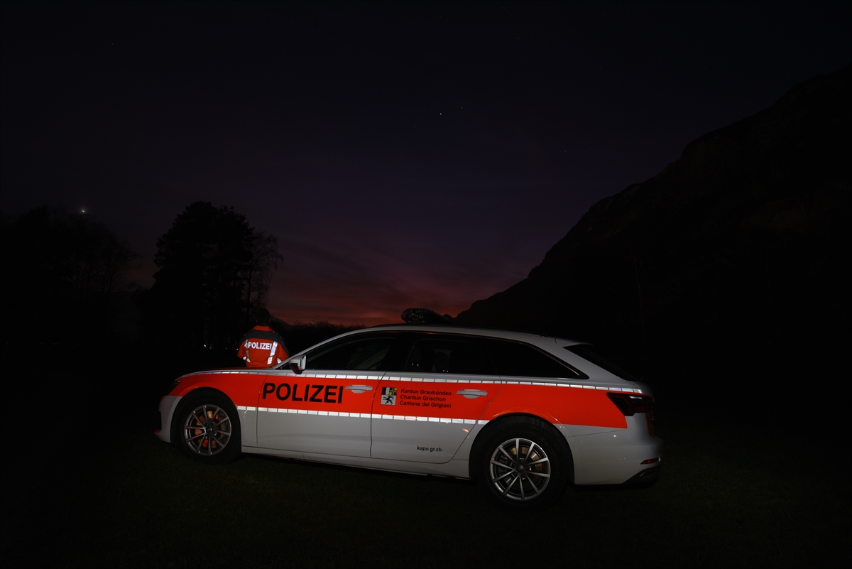 Nachtaufnahme. Im Vordergrund ein angeleuchteter Patrouillenwagen, links davon leicht im Hintergrund ein Polizist. Im Hintergrund zeichnen sich Berge vom Horizont ab.
