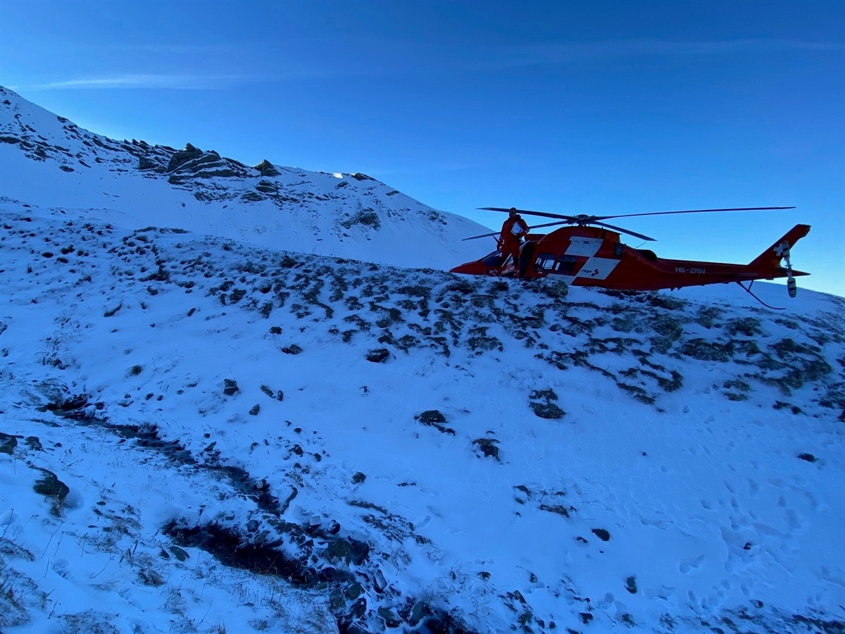 Ein ansteigender schneebedeckter Hang. Auf einer Anhöhe ein Rega-Helikopter, vor diesem ein Crew-Mitglied. Im Hintergrund blauer Himmel.