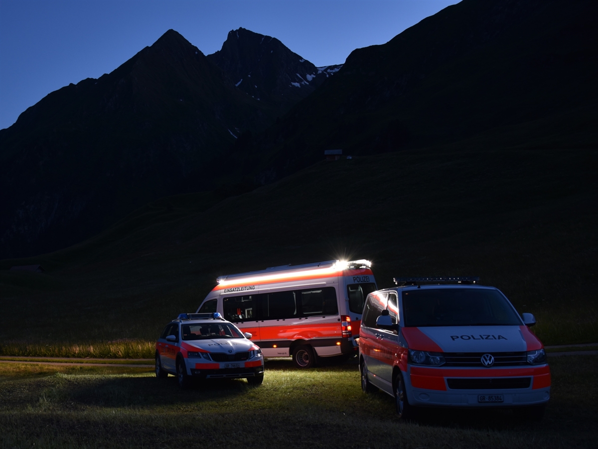 Einsatzfahrzeuge der Kantonspolizei Graubünden auf einer Wiese im Gebirge in der Nachtdämmerung.