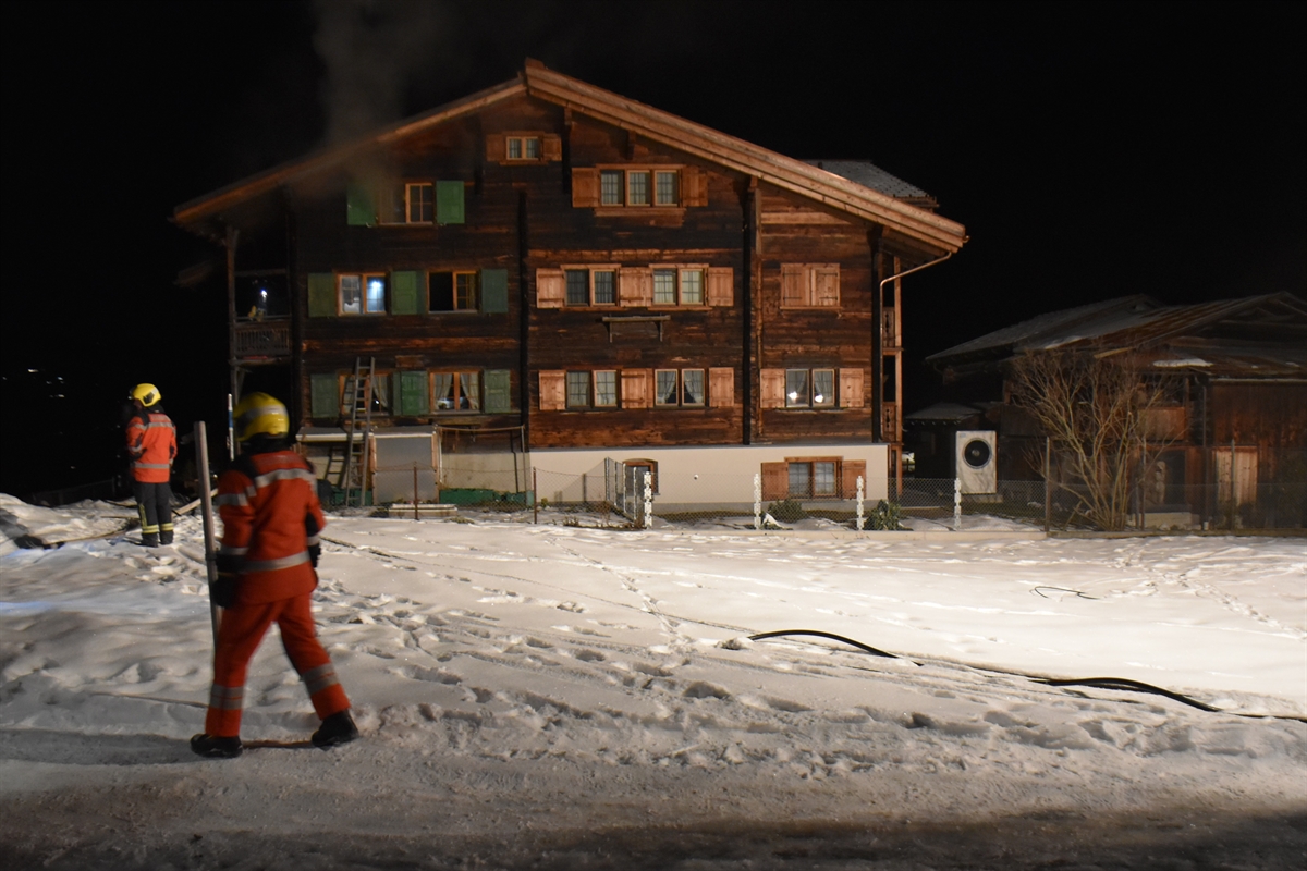 Nachtaufnahme mit links 2 Feuerwehrleuten im Vordergrund, einer verschneiten Wiese und im Hintergrund das 4-stöckige Holzhaus, in welchem es brannte. Dort angelehnt eine Leiter. Aus einem geöffneten Fenster steigt Rauch auf.
