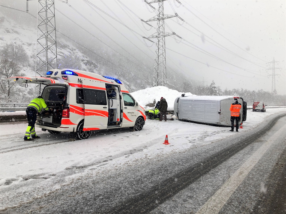Blick auf die A13. Links ein Ambulanzfahrzeug, rechts davon der auf der Fahrerseite liegende Lieferwagen. Das Ambulanzteam wird unterstützt von zwei weiteren Personen. Ein Polizist leitet den Einsatz.
