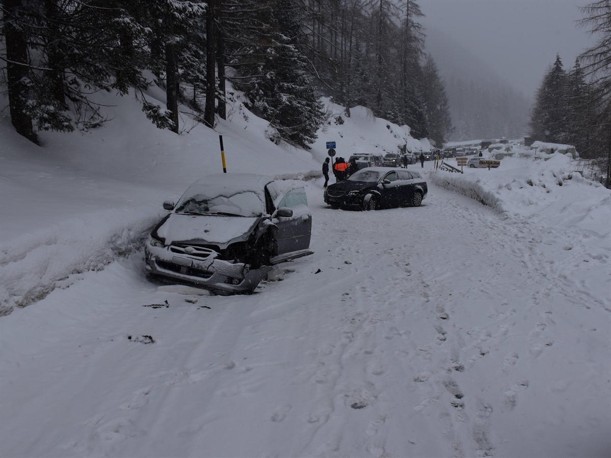 Winterliche Strasse, links von vorne das frontal-seitlich stark beschädigte bergwärts gefahrene Auto. Dahinter das andere Auto, ebenfalls mit stark beschädigter Front. Im Hintergrund Personen.
