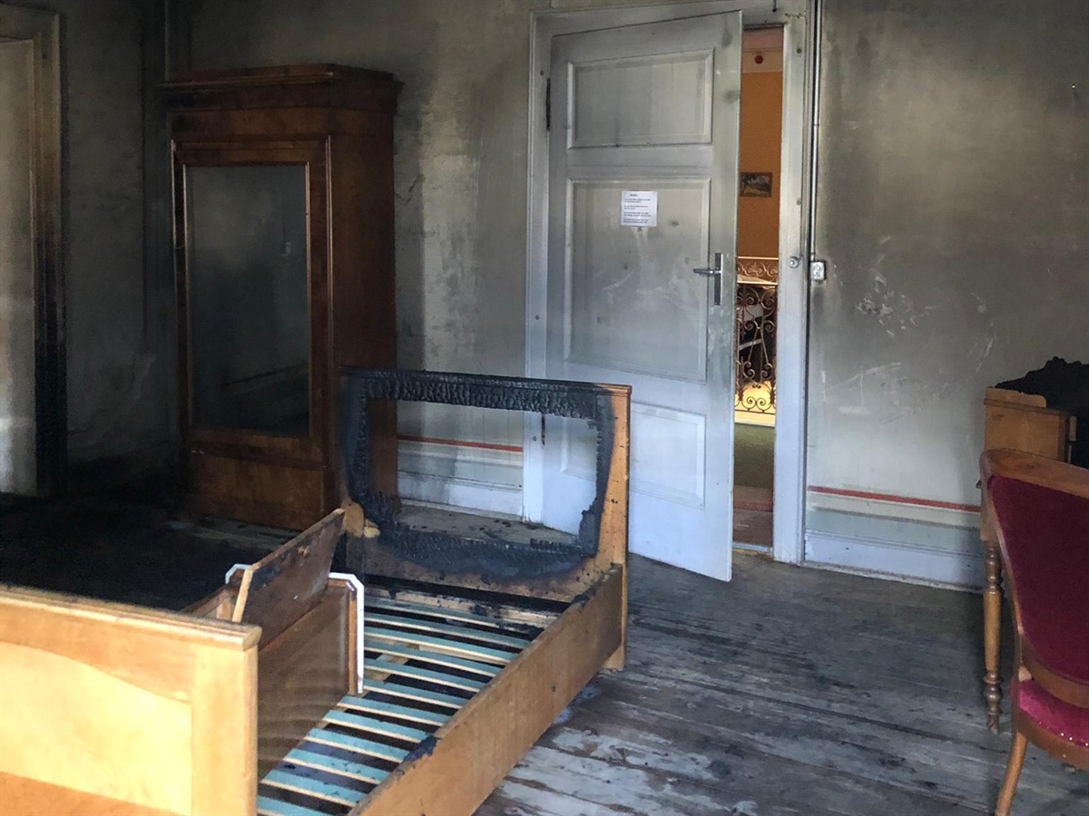 Im Brandzimmer steht ein verkohltes Bett. Durch einen Türspalt sieht man ins Treppenhaus. Überall ist Rauchschaden erkennbar. 