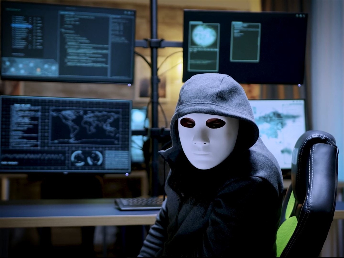 Im Hintergrund Bildschirme. Im Vordergrund eine maskierte Person auf einem Bürostuhl mit frontalem Blick zur Kamera. 