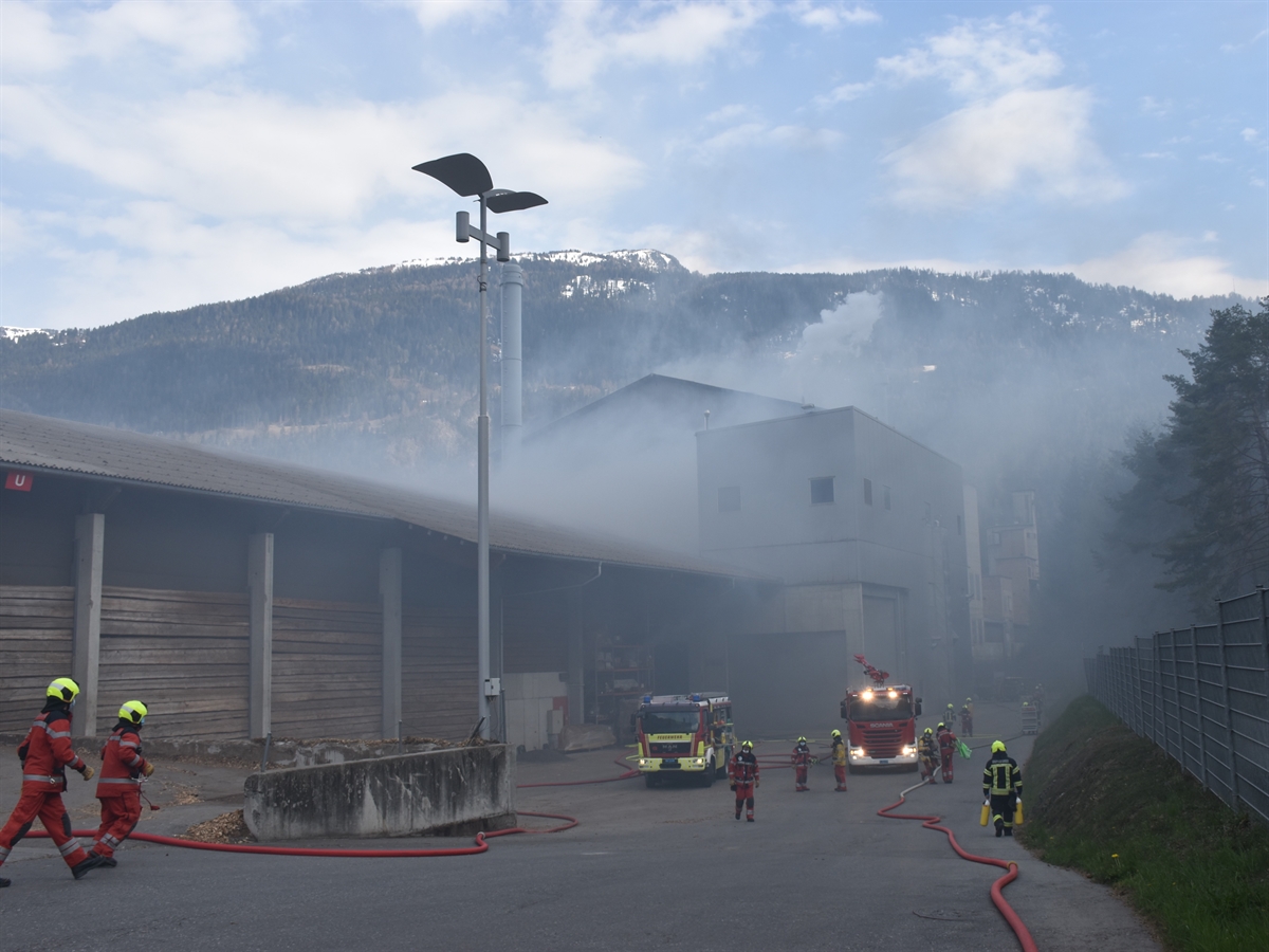 Der Brandort von aussen, Rauch dringt aus zwei Gebäuden. Zwei Feuerwehrautos und mehrere Feuerwehrleute im Einsatz.