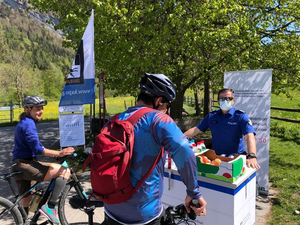 Links eine Velofahrerin, halbwegs auf dem Sattel steht sie mit einem weiteren Velofahrer am Präventionsstand. Im Hintergrund Landschaft.