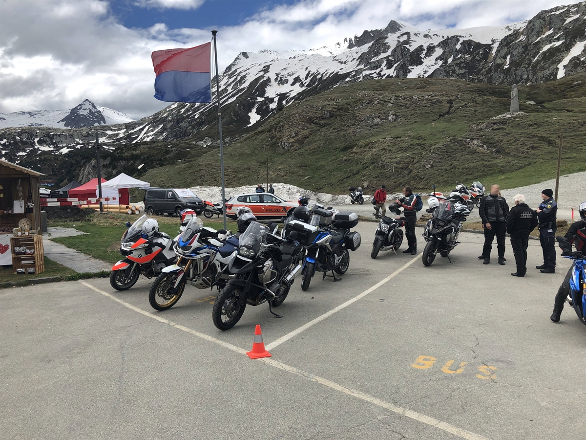 Ein Parkplatz mit mehreren Motorrädern – darunter eines der Polizei – und Motorradfahrern. In Bei einer Gruppierung ein Polizist. Im Hintergrund Bergpanorama.
