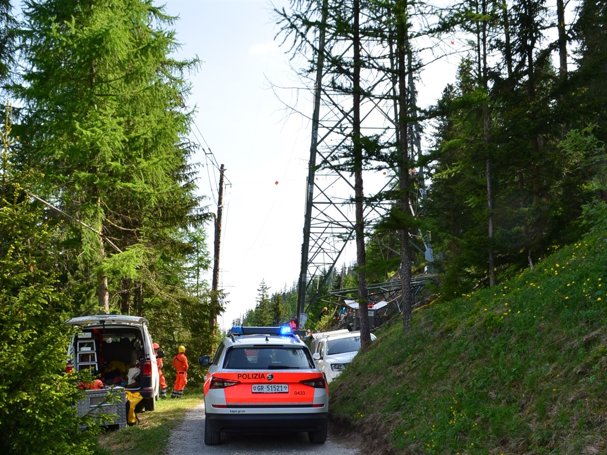 Hochspannungsmast am Rande eines Waldweges. Auf dem Weg steht ein Patrouillenfahrzeug der Kantonspolizei Graubünden sowie zwei Fahrzeuge der Freileitungsfirma. Zudem stehen mehrere Arbeiter neben, unter sowie um den Hochspannungsmast.