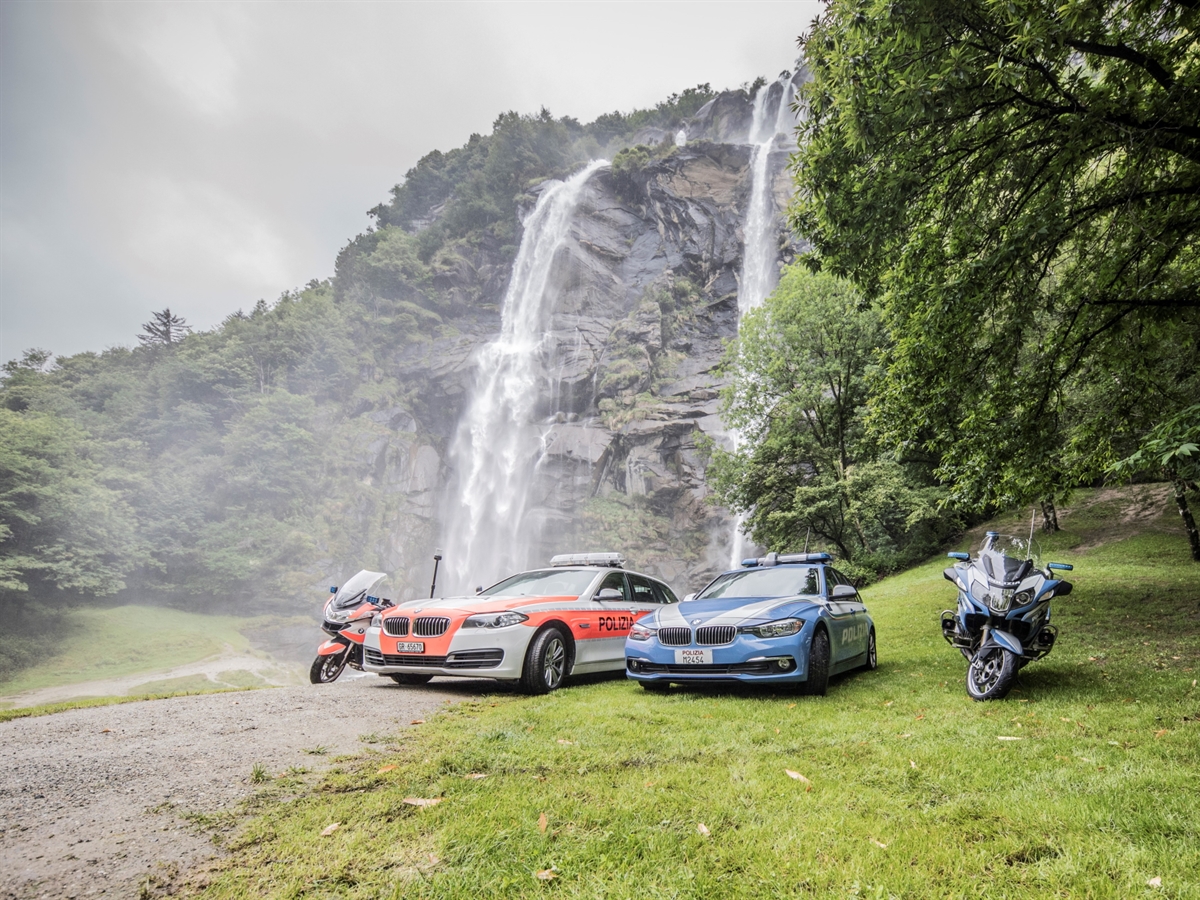 Ein Bündner und ein Italienisches Patrouillenfahrzeug posieren mit je einem Motorrad vor einem malerischen Wasserfall.