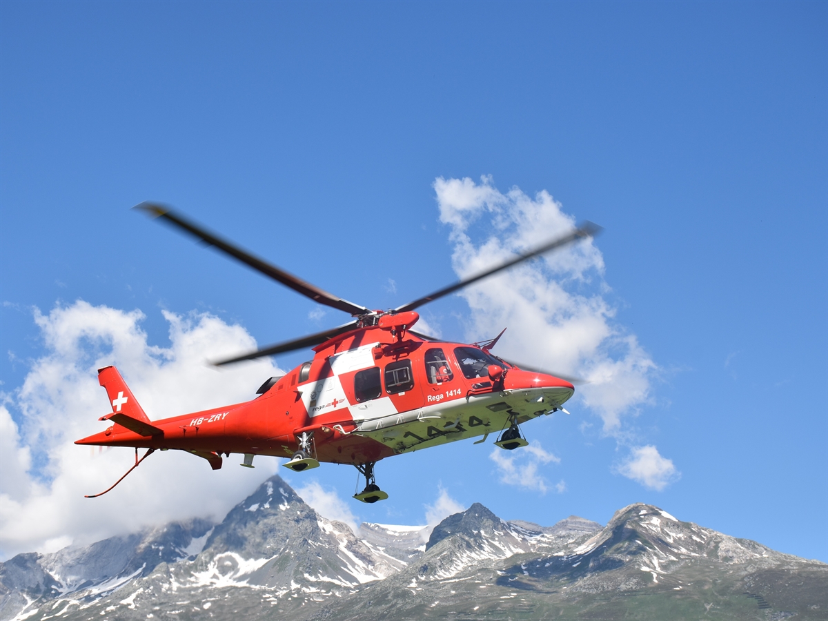 Ein Helikopter der Rega bei bestem Wetter vor einer eindrücklichen Bergkette im Kanton Graubünden