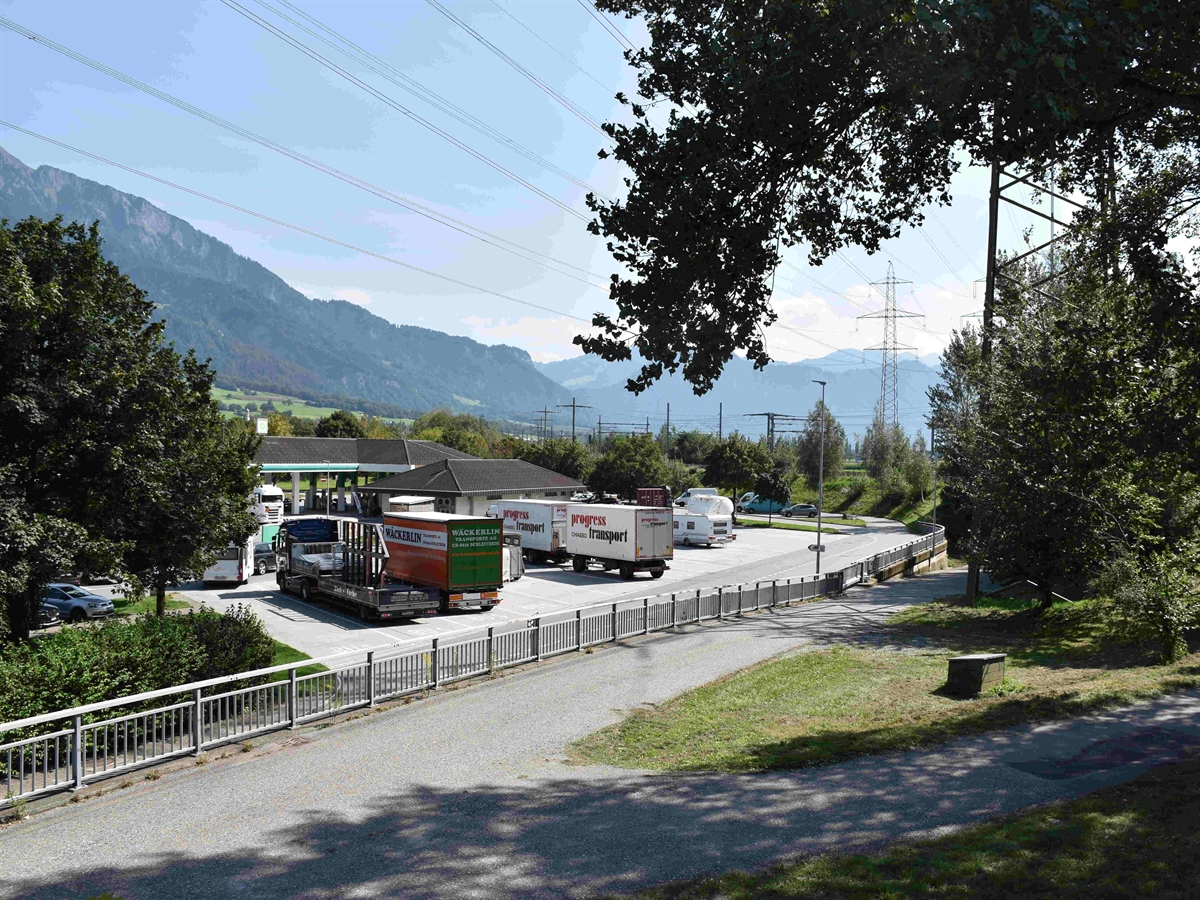 Der Parkplatz auf welchem diverse Fahrzeuge stehen. Im Vordergrund eine Verbindungsstrasse, im Hintergrund Berge und Horizont.