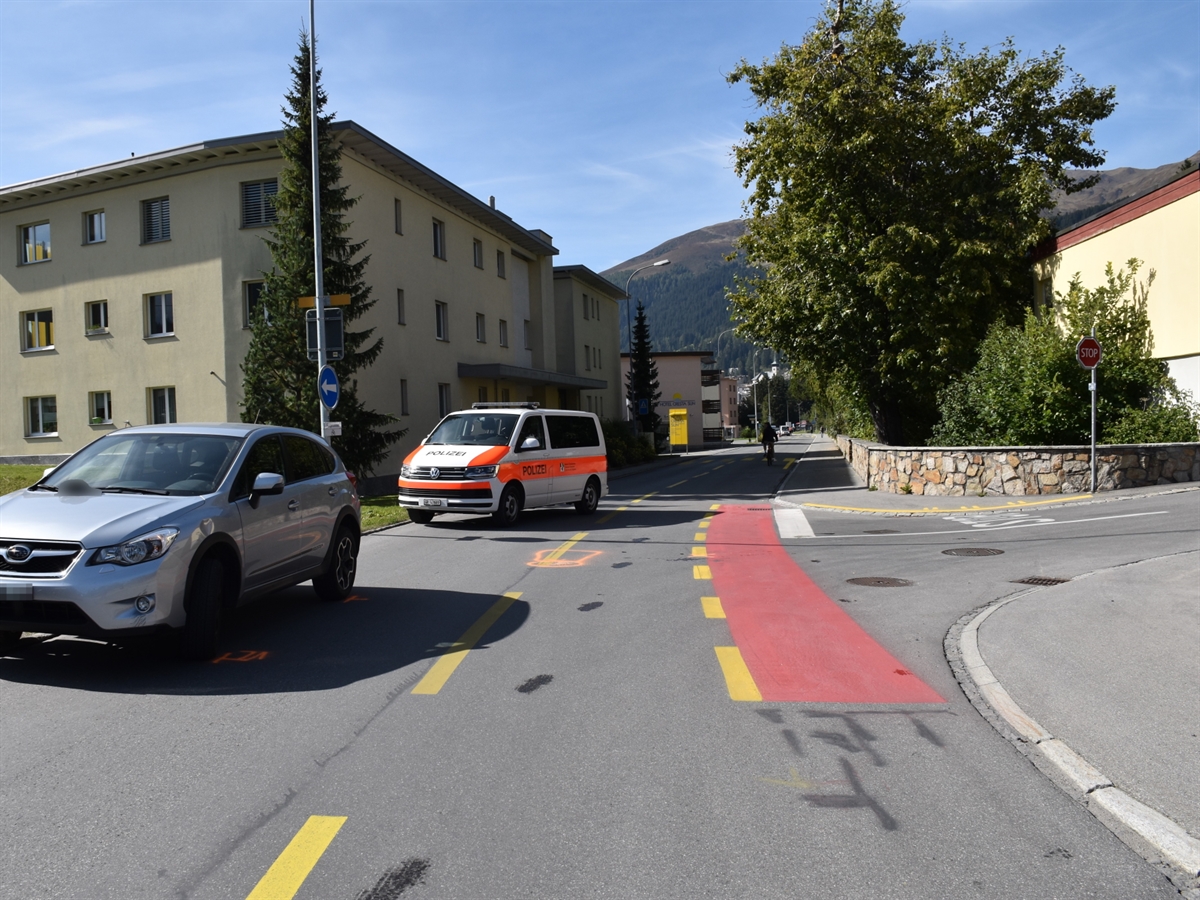 Talstrasse in Richtung Bahnhof Davos Platz gesehen. Rechts die Einmündung mit dem Stop auf dem Reginaweg. Auf der Talstrasse ist rot der Fahrradstreifen markiert. Links im Bild das am Unfall beteiligte Auto. Im Hintergrund ein Patrouillenfahrzeug der Kapo GR. Das farbenfrohe Bild wird mit blauem Himmel ergänzt.