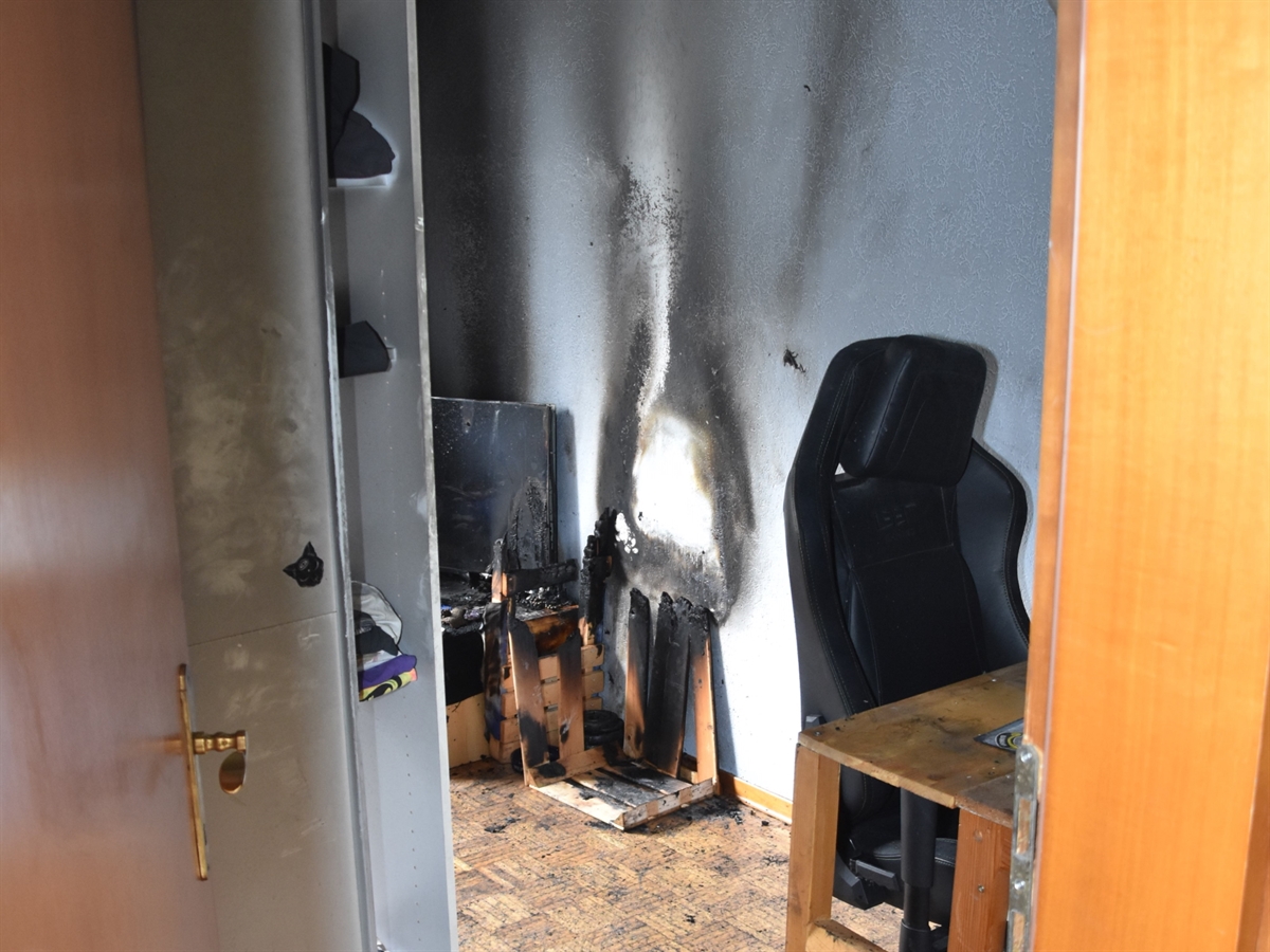 Blick durch die Türe in das Zimmer. In diesem ist eine angesengte Wand, ein verbranntes Holzgestell sowie ein durch den Brand beschädigter Fernseher sichtbar.