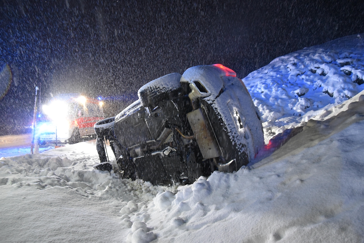 Im Vordergrund der auf der rechten Seite liegende beschädigte Personenwagen im Schnee. Im linken Hintergrund ist in der Dunkelheit im Schneetreiben ein Patrouillenfahrzeug der Polizei zu erkennen.