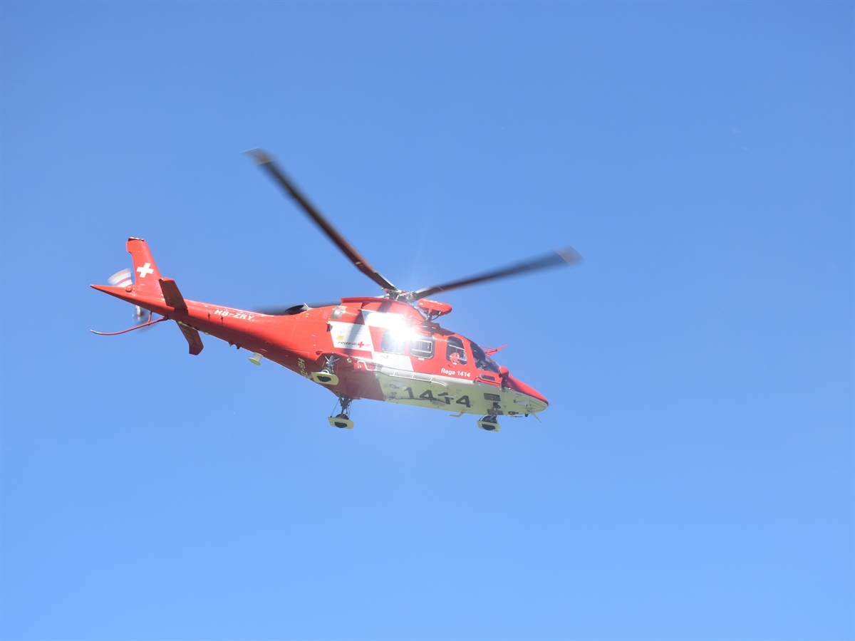 Ein fliegender rot-weisser Helikopter der Rega welcher sich von schräg unten gegen den blauen Himmel abgrenzt.