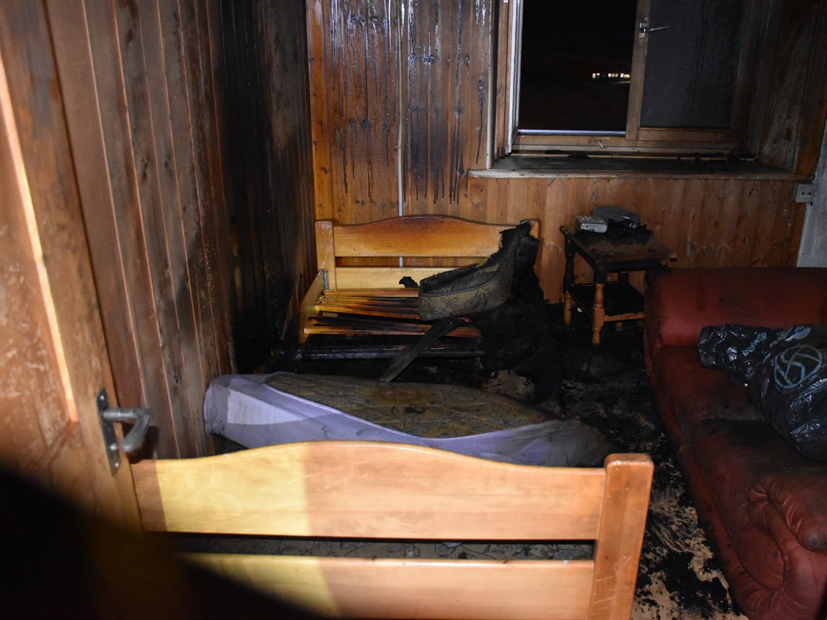 Ein Zimmer mit diversen angebrannten Gegenstände. Ein Fenster ist offen, an den mit Holz getäferten Wänden sind Brandspuren erkennbar. 