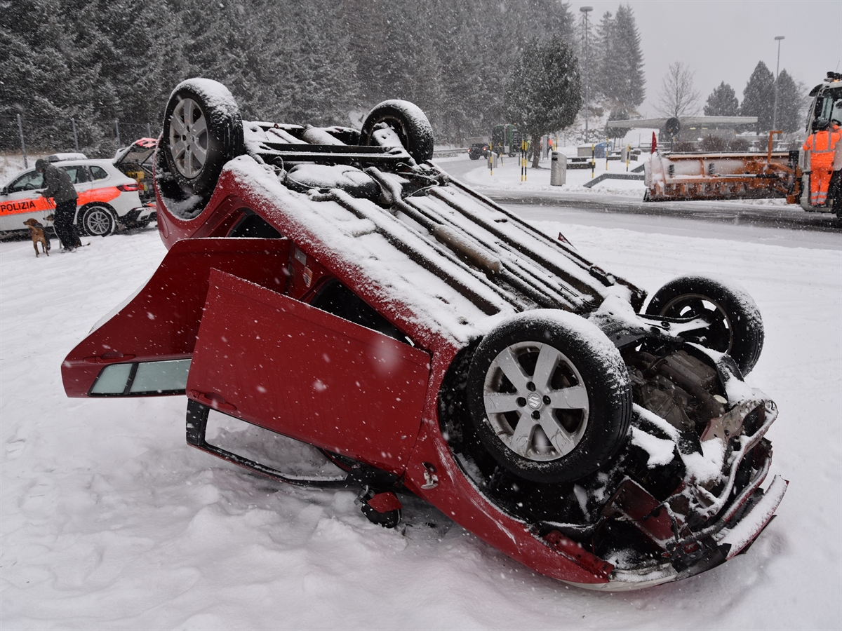 Ein roter auf dem Dach liegender Kleinwagen. Die Umgebung ist verschneit. Links im Hintergrund ein Polizeiauto, rechts im Hintergrund ein Lastwagen mit Schneepflug.