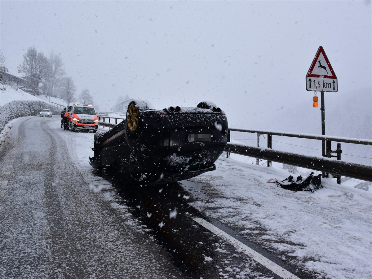 Ein schwarzes, inmitten der Strasse, auf dem Dach Auto liegendes Auto. Die Umgebung ist verschneit. Links im Hintergrund ein weiter oben an der Steigung stehendes Polizeiauto. 