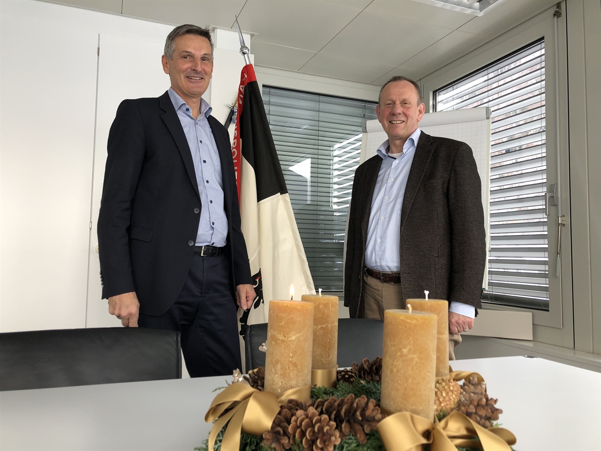 Walter Schlegel und Bruno Zanga bei der Geschäftsübergabe am 29. November 2021 in Chur. In der Mitte die Korpsfahne der Kantonspolizei Graubünden und im vordergrund auf dem Tisch ein Adventskranz mit einer brennenden Kerze.
