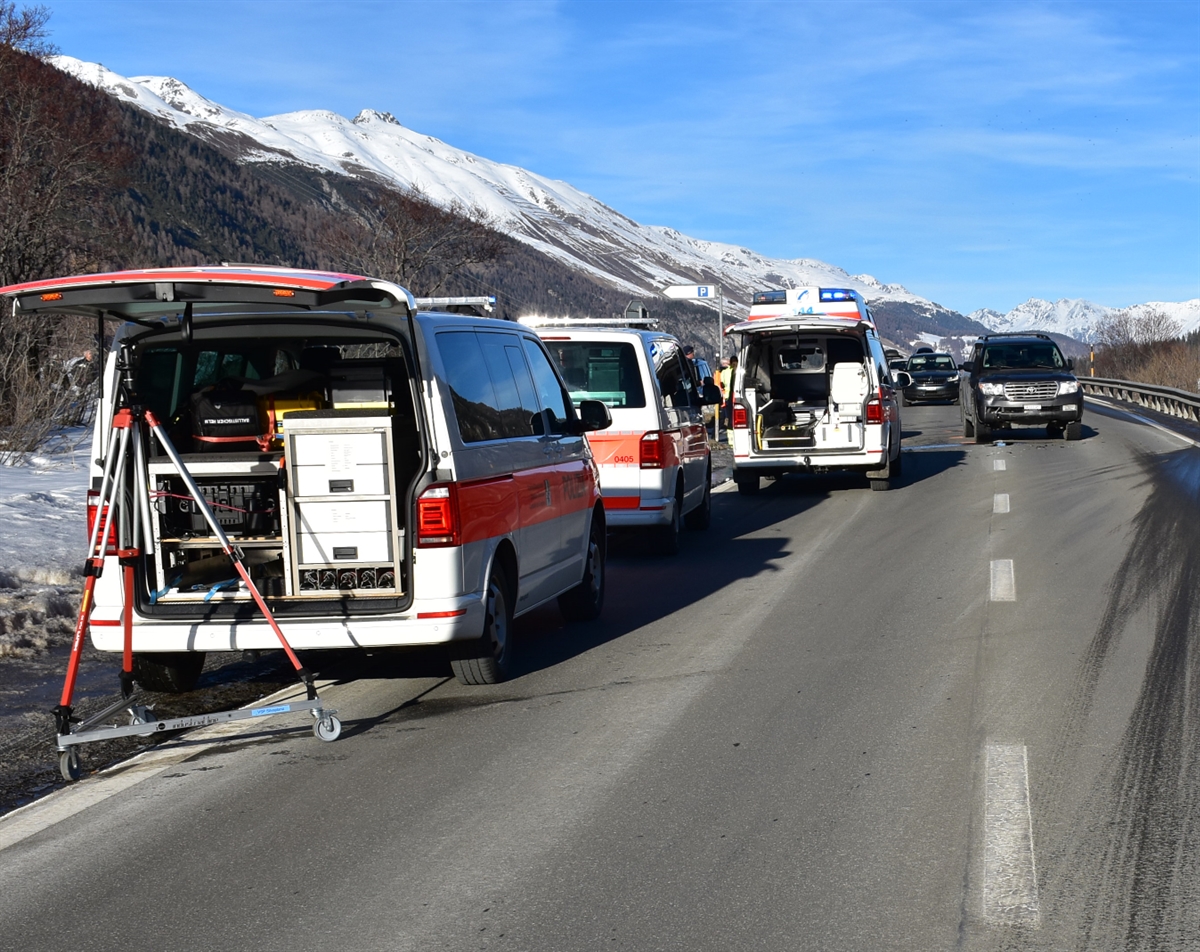 Im Hintergrund sind verschneite Berge zu erkennen. Es ist blauer Himmel. Auf der linken Seite stehen am Strassenrand drei Einsatzfahrzeuge der Rettungskräfte und der Polizei. Im Hintergrund ist ein am Unfall beteiligtes Fahrzeug zu erkennen.