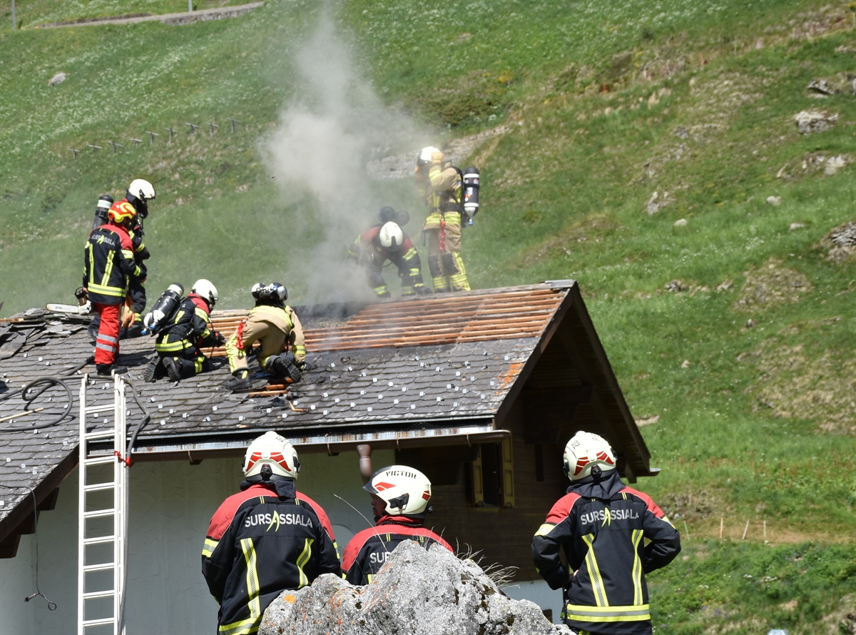 Im Vordergrund stehen drei Feuerwehrleute. Im Hintergrund das Einfamilienhaus. Es ist zu erkennen wie der Dachstock raucht. Zudem sind noch weitere Feuerwehrleute auf dem Dach zu erkennen, welche den Brand bekämpfen.
