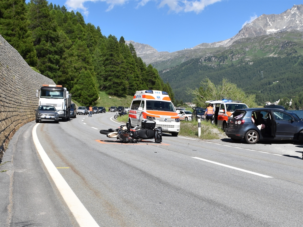 Motorrad liegt auf der Malojastrasse. Rechts davon das Auto auf welches der Motorradfahrer aufgefahren ist. Hinter dem Motorrad ein Ambulanzfahrzeug und daneben ein Patrouillenfahrzeug der Kantonspolizei Graubünden. Blauer Himmel rundet das Bild ab.