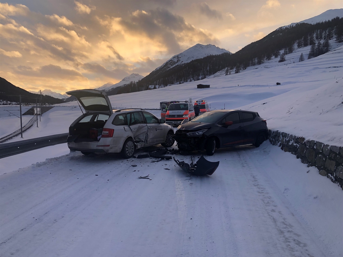 Zwei Autos, sie stehen mit der Front dicht einander zugewendet, die die Strasse blockieren. Weiter entfernt ein Polizei- und ein Ambulanzfahrzeug. Im Hintergrund Berge und stimmungsvoller Dämmerungshimmel.