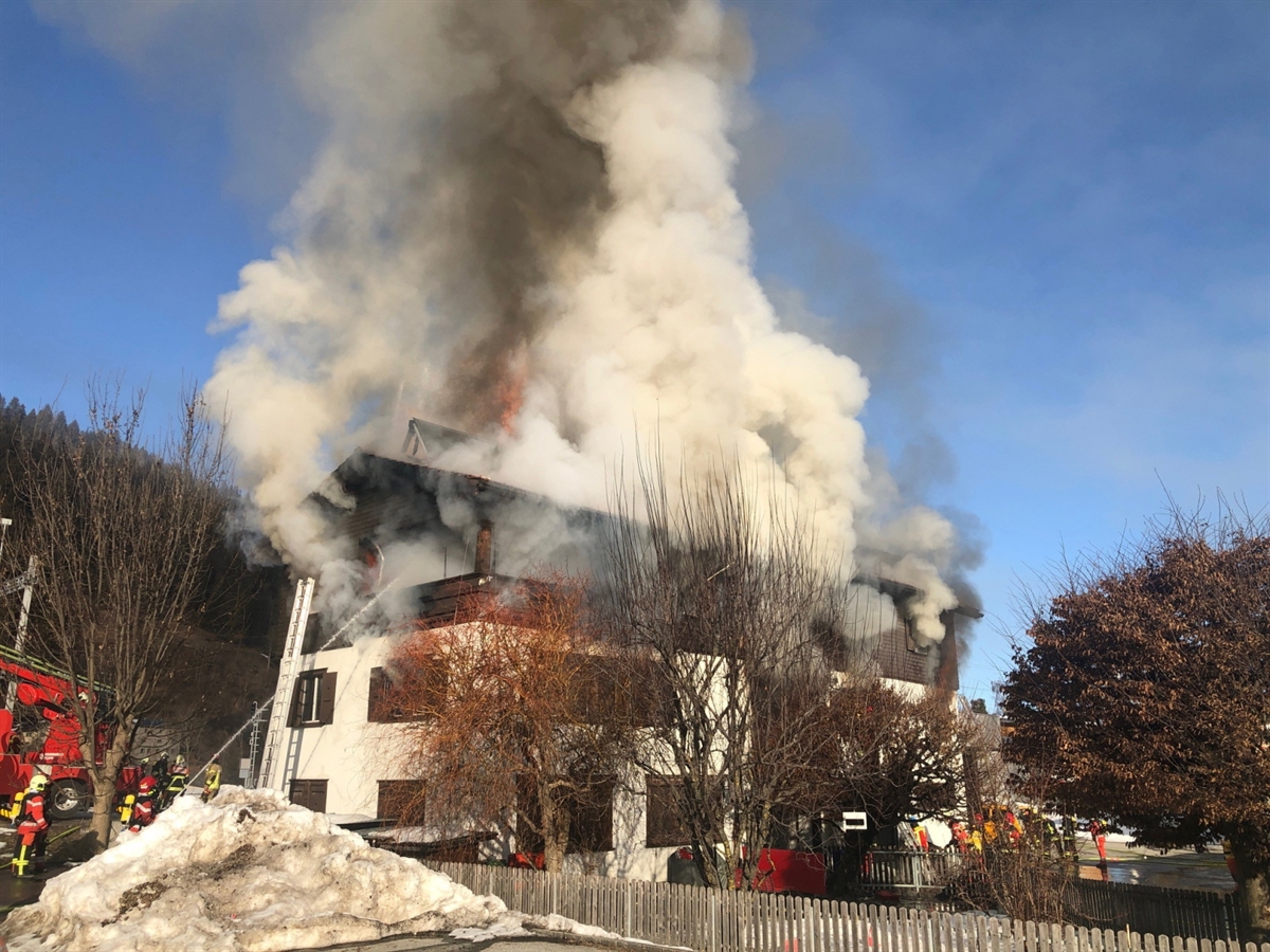 Aus dem dreigeschossigen Wohnhaus tritt starker Rauch heraus und aus dem Dachstock sind Flammen sichtbar. Rund um das Haus sind Feuerwehrleute bei den Löscharbeiten.
