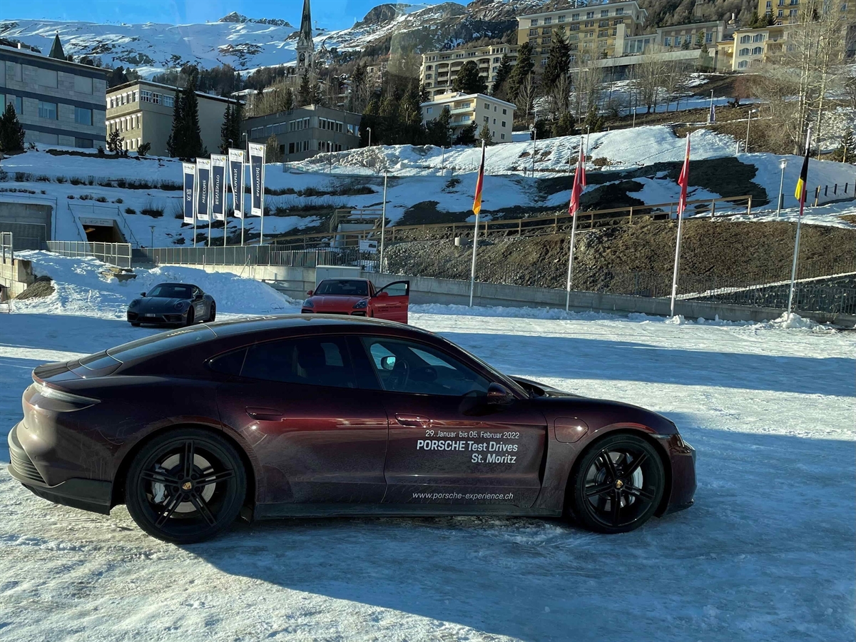 Ein auf einem schneebedeckten Parkplatz abgestellter roter Porsche Taycan. Im Hintergrund zwei weitere Autos für Probefahrten. Im Hintergrund St. Moritz Dorf und das Skigebiet. Am Horizont blauer Himmel.