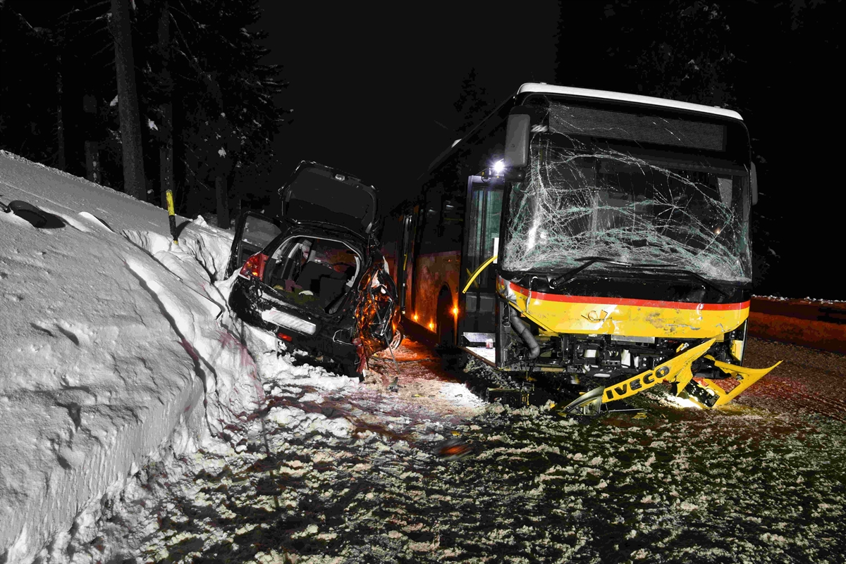 Ein Postauto mit Frontbeschädigung auf der schneebedeckten Fahrbahn und ein beschädigter Personenwagen mit Heckansicht am Fahrbahnrand im Schnee zwischen Postauto und Böschung.