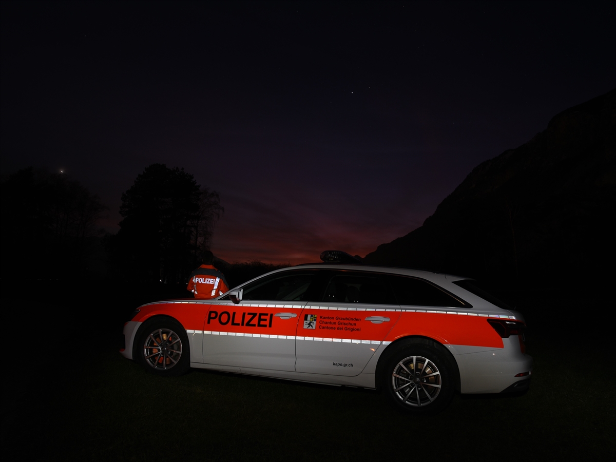 Nachtaufnahme mit Blick auf die linke Seite eines Polizeiautos. Links hinter dem Auto ein Polizist. In der Ferne Berge in der Dämmerung.