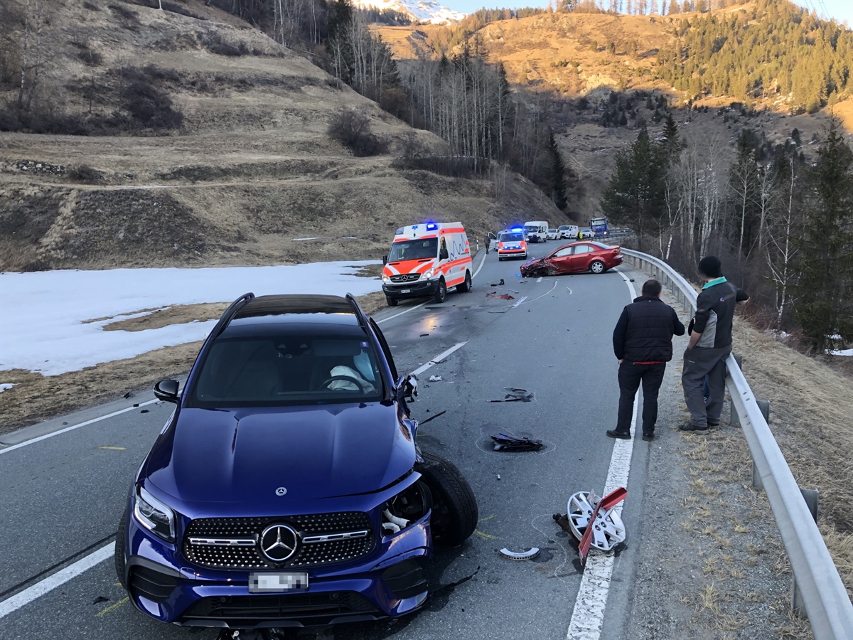 Im Vordergrund ein blaues stark beschädigtes Auto. Im Hintergrund ein rotes ebenfalls stark beschädigtes Auto. Weiter ein Ambulanzfahrzeug, ein Polizeiauto und Kollisionsspuren.