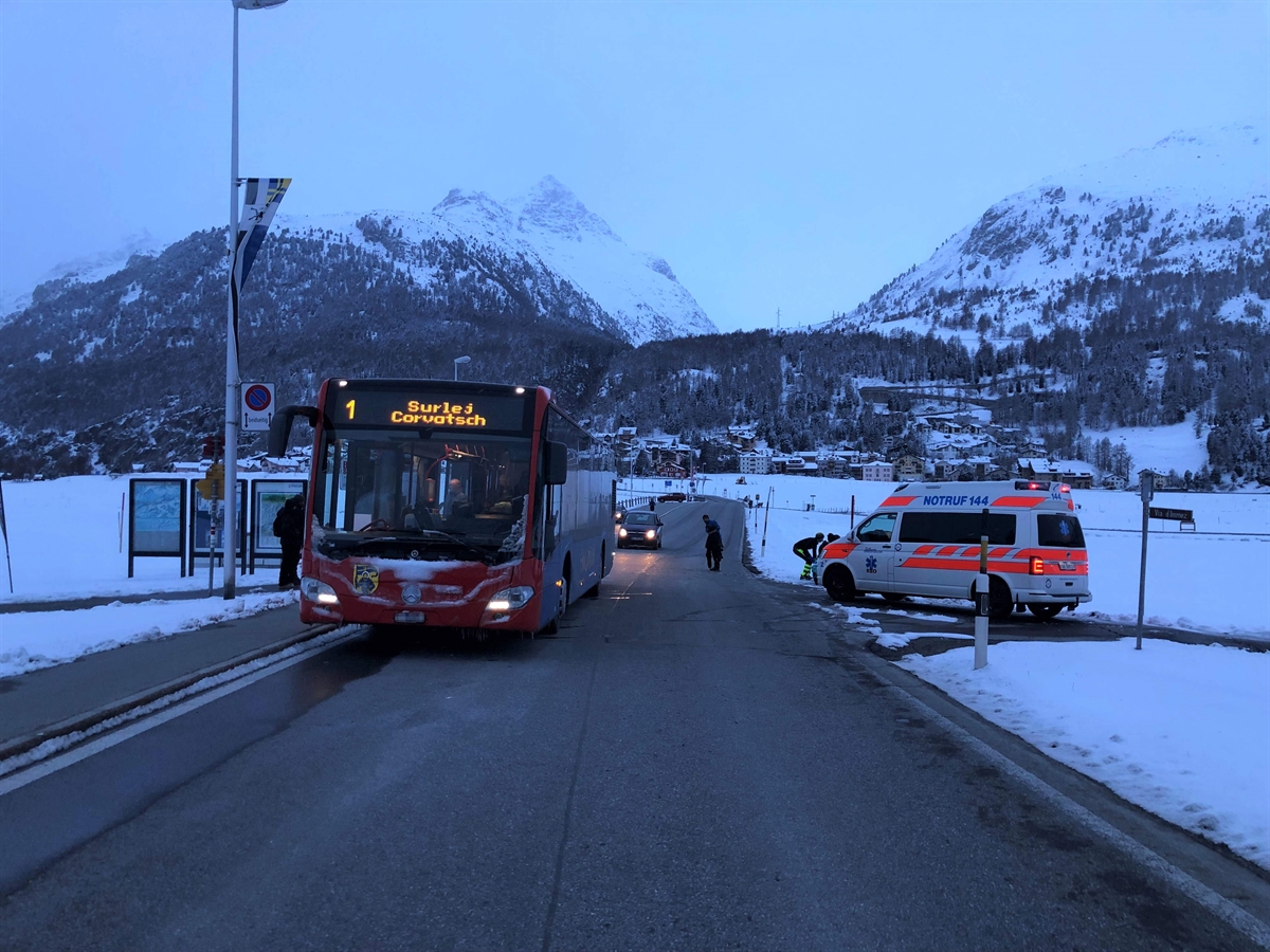 Die schwarz geräumte Strasse, ringsum Winterlandschaft. Links auf der Strasse von vorne ein Linienbus, rechts in einer Einmündung ein Ambulanzfahrzeug. Im Hintergrund Berge.