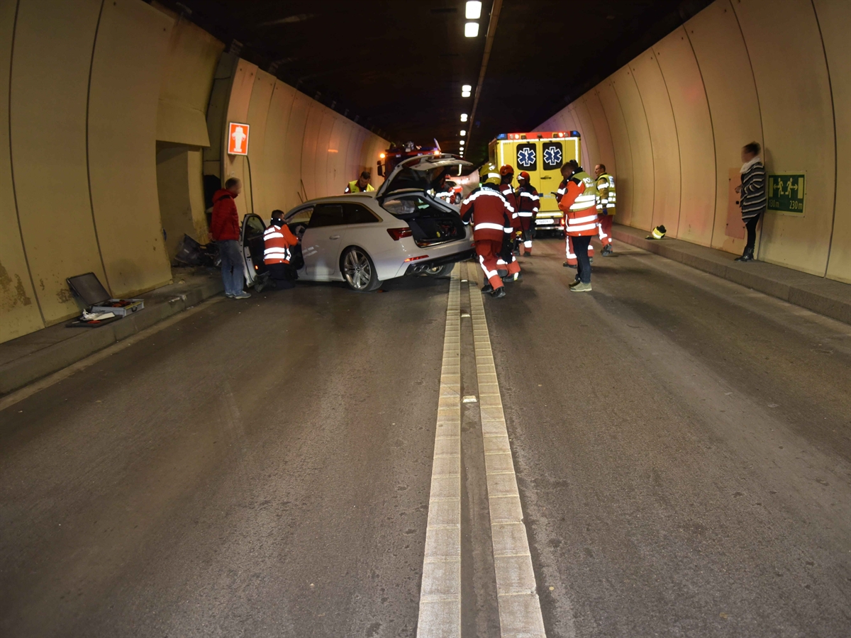 Links das frontal total beschädigte Auto an der Tunnelwand, umgeben von Rettungskräften. Im Hintergrund ein Ambulanz- sowie ein Feuerwehrfahrzeug.